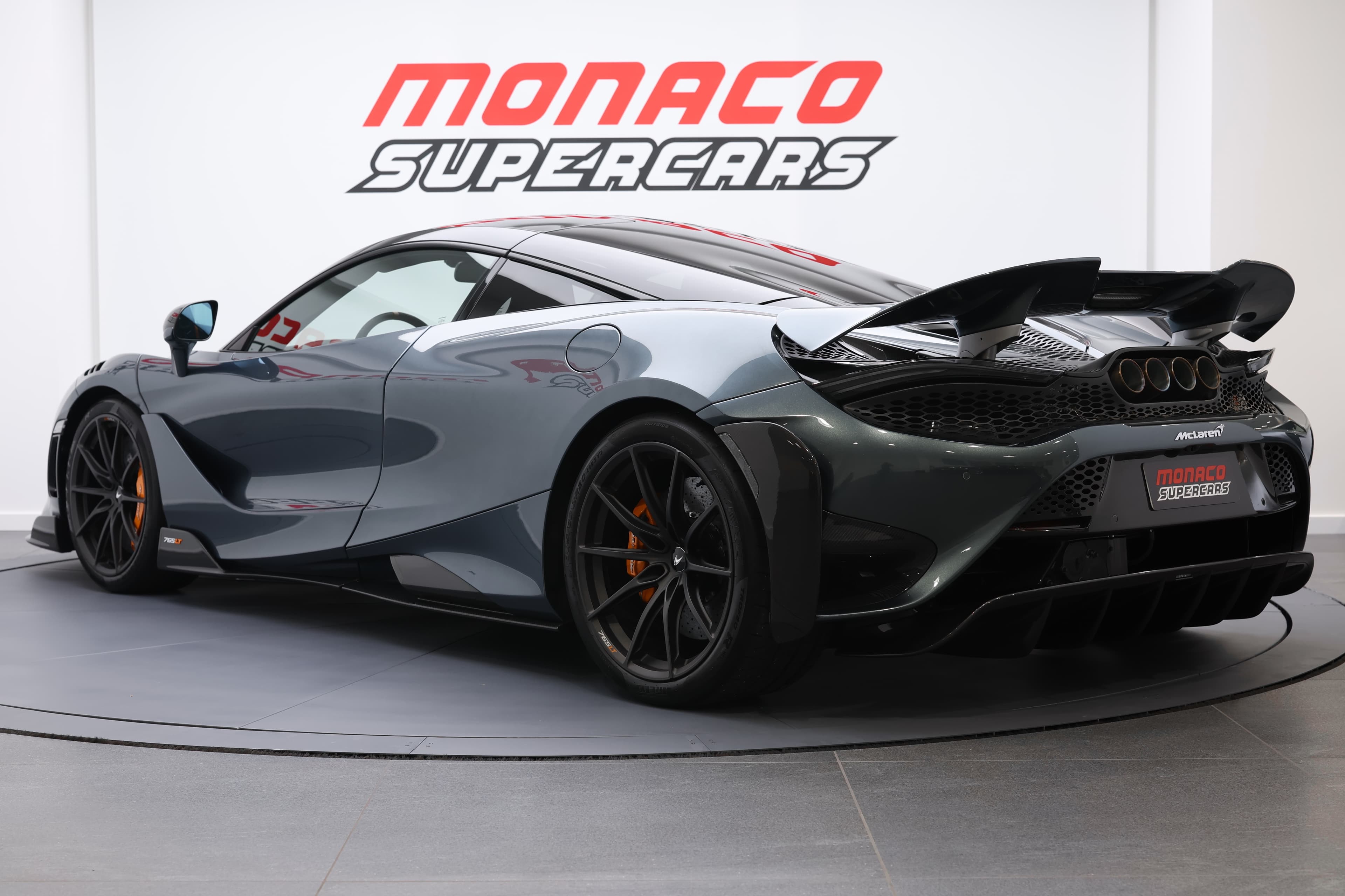 L'image montre une McLaren 765LT vue en 3/4 arrière, mettant en avant ses courbes dynamiques, ses feux arrière distinctifs et l'aérodynamisme de sa carrosserie, soulignant son allure sportive et son design audacieux.