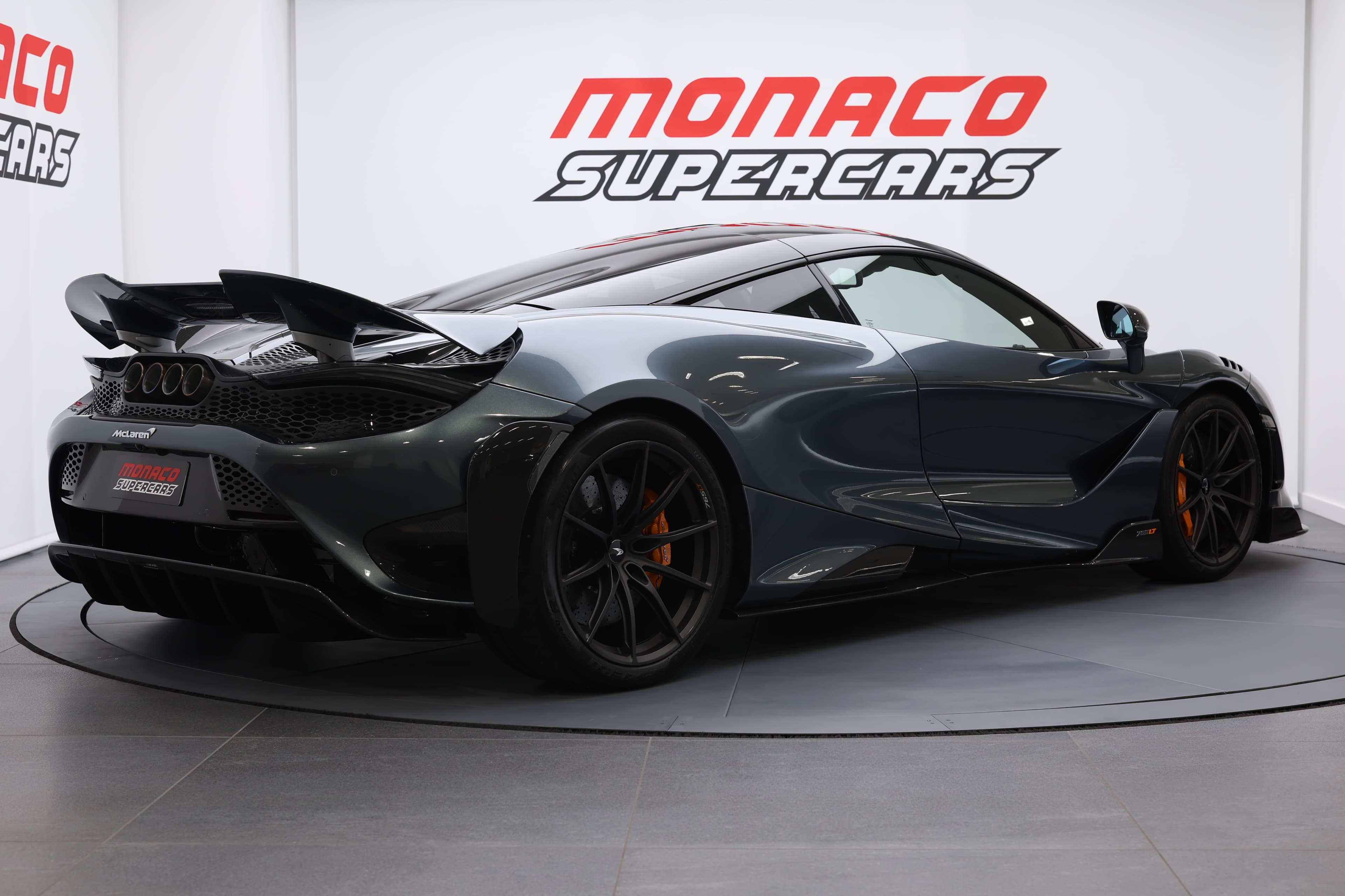L'image montre une McLaren 765LT vue en 3/4 arrière, mettant en avant ses courbes dynamiques, ses feux arrière distinctifs et l'aérodynamisme de sa carrosserie, soulignant son allure sportive et son design audacieux.