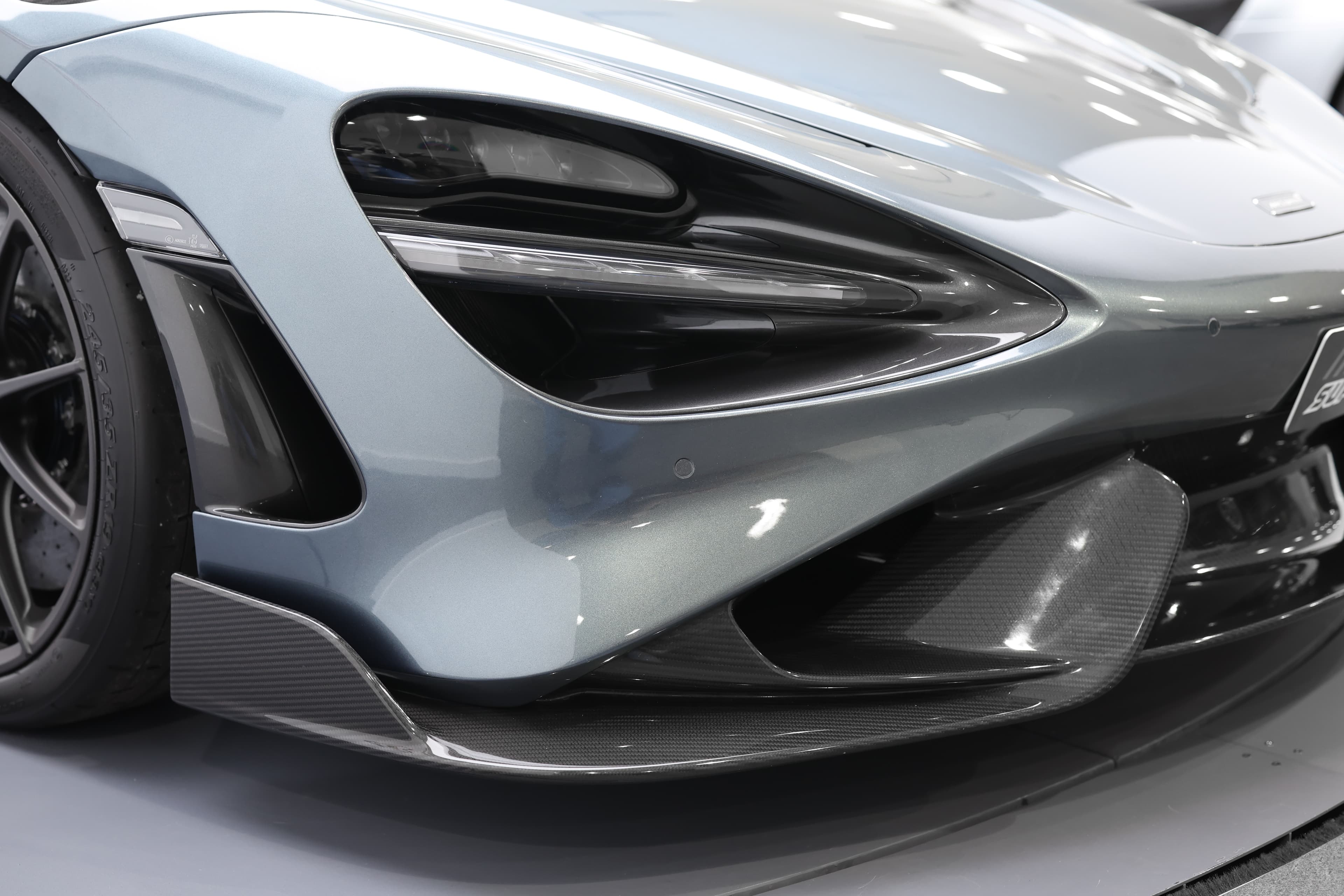 L'image se concentre sur le feu avant d'une McLaren 765LT, mettant en valeur son design moderne et angulaire, ainsi que la technologie d'éclairage sophistiquée qui accentue l'esthétique agressive et dynamique de la voiture.