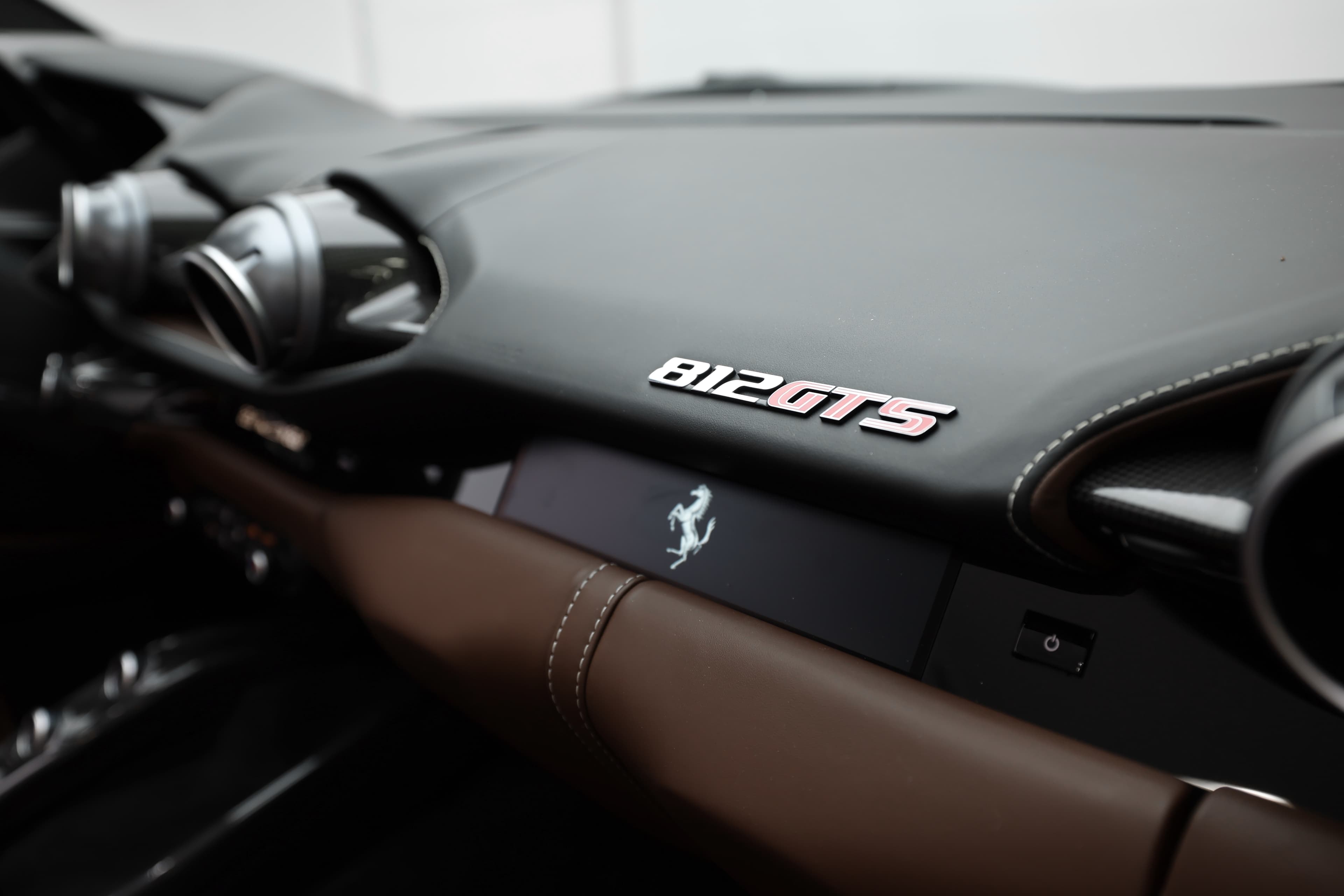 "Regardez de plus près le tableau de bord de la Ferrari 812 GTS, mettant en valeur le badge distinctif et les matériaux haut de gamme. L'alliance du cuir chocolat et des éléments en carbone souligne le luxe et la performance de ce modèle emblématique."