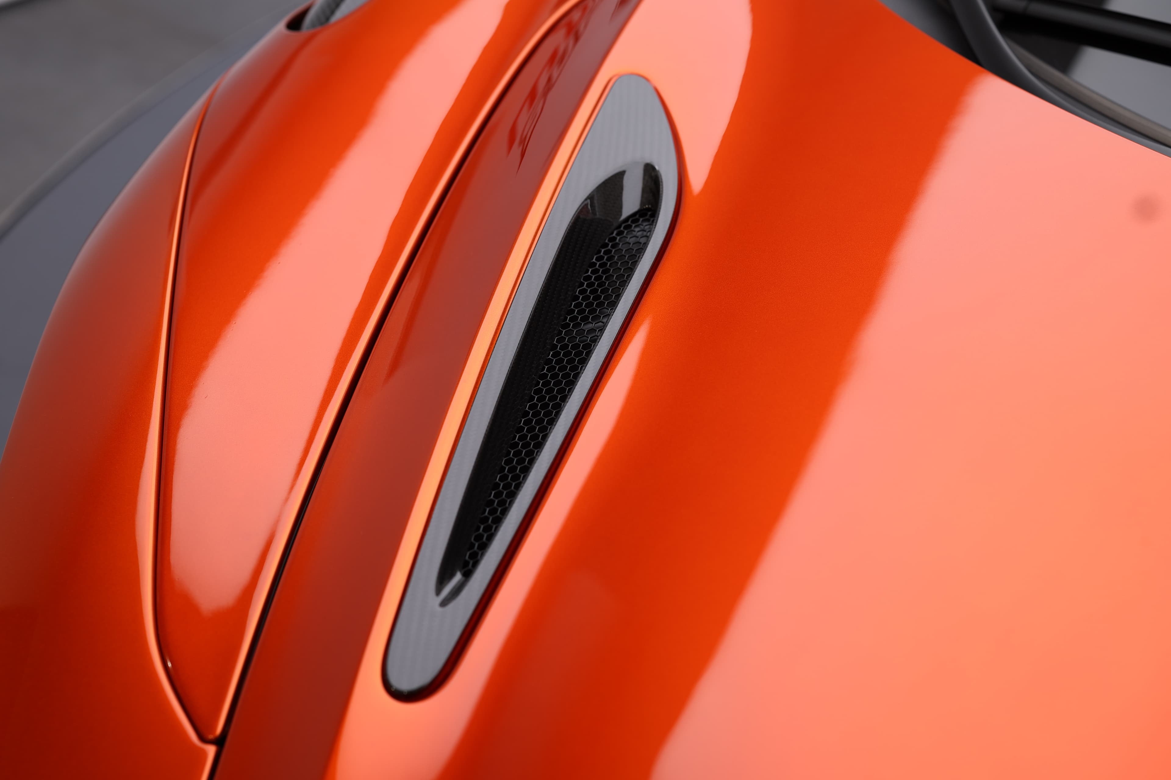 Gros plan sur une prise d'air de la McLaren 765LT volcano orange, mettant en évidence le contraste entre la peinture brillante et le matériau en carbone, illustrant l'importance de l'aérodynamisme et du design dans cette supercar.