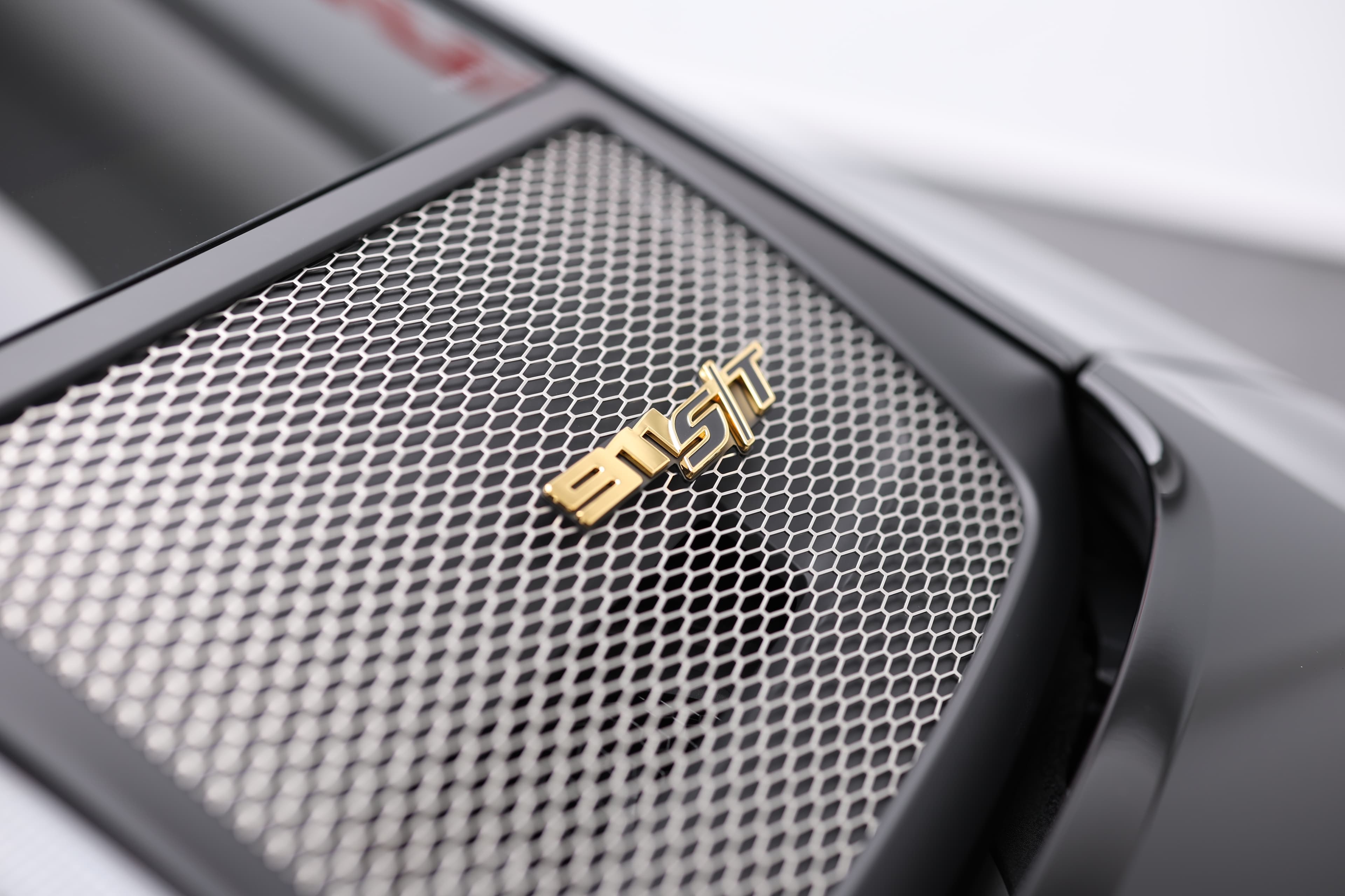 Gros plan sur la grille du capot arrière de la Porsche 911, mettant en avant le logo "S/T" en doré, qui souligne le caractère exclusif et sportif du véhicule. Ce détail raffiné témoigne de l'attention portée à la performance et au design dans chaque aspect de la voiture.